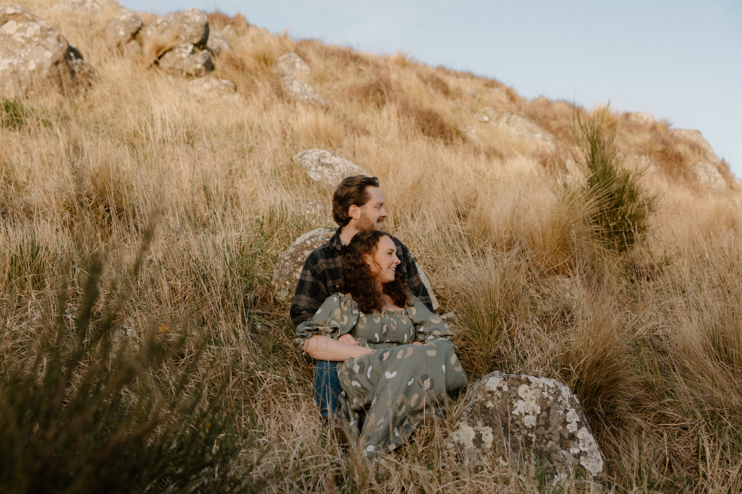 Couple sitting in the grass during an engagement session at Rapaki Rock in the Port Hills, Christchurch.
