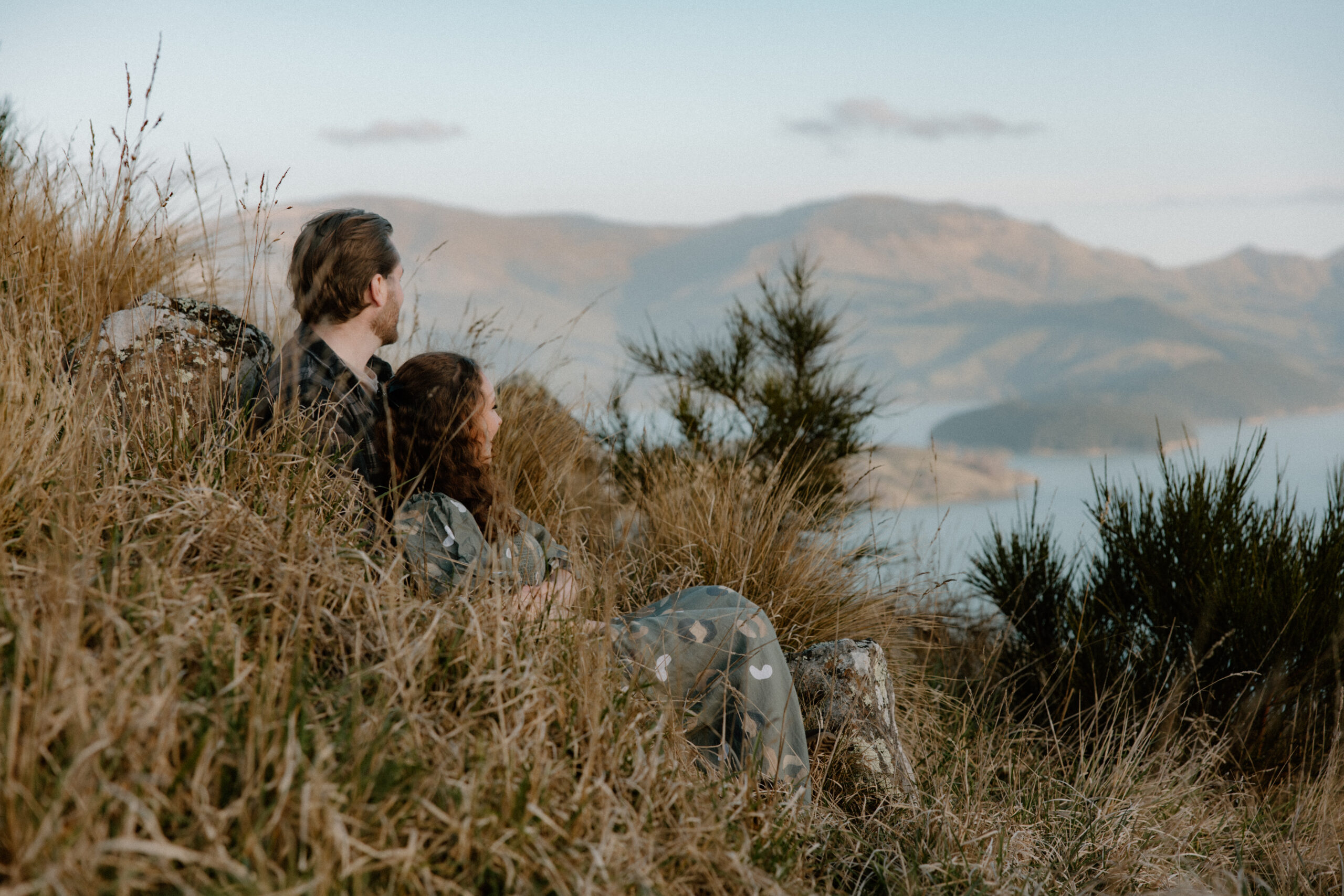Couple sitting in the grass during an engagement session at Rapaki Rock in the Port Hills, Christchurch.