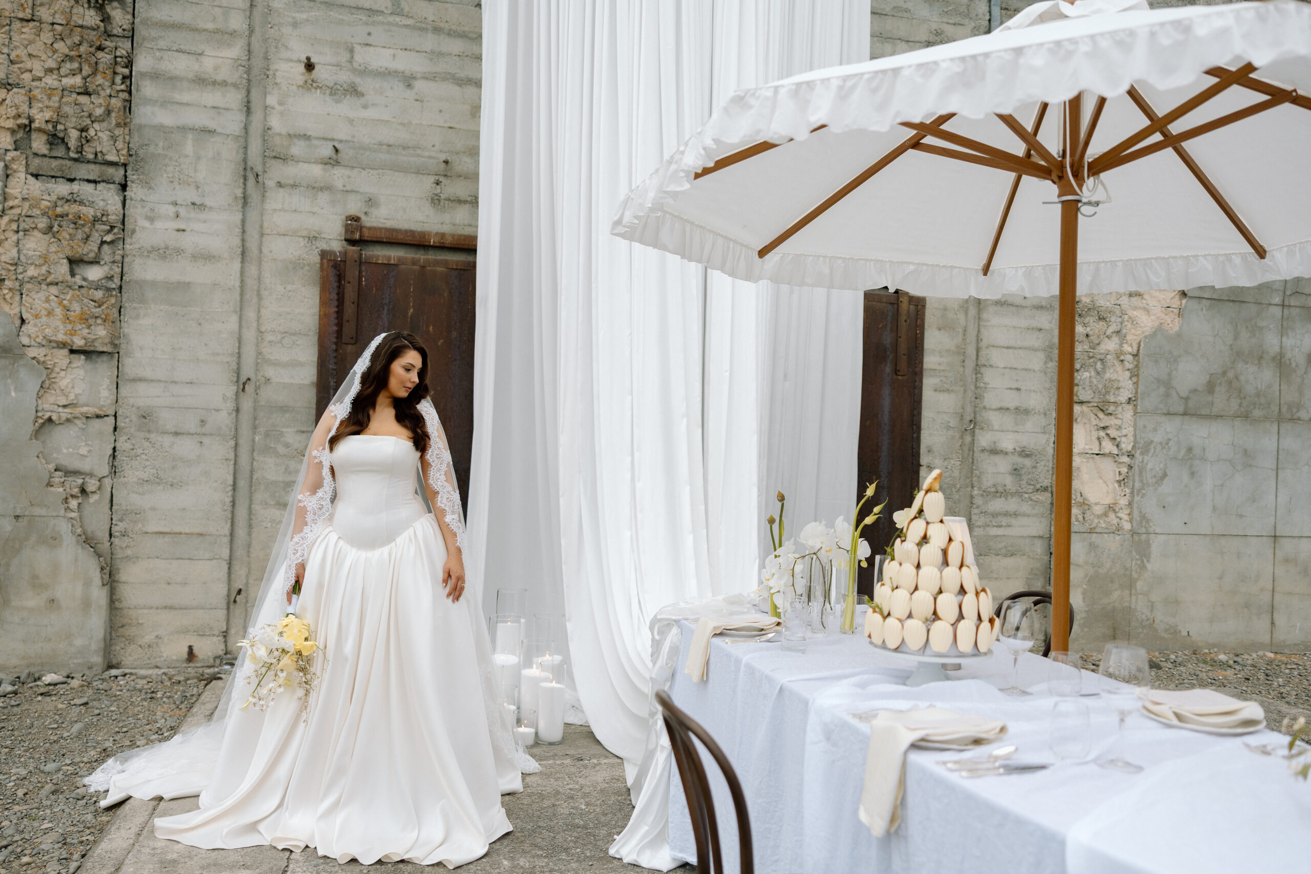 Bride in a strapless satin corset gown with a drop-waist skirt standing behind a styled wedding table, beneath a white ruffled umbrella at The Flaxmill in Oxford, New Zealand