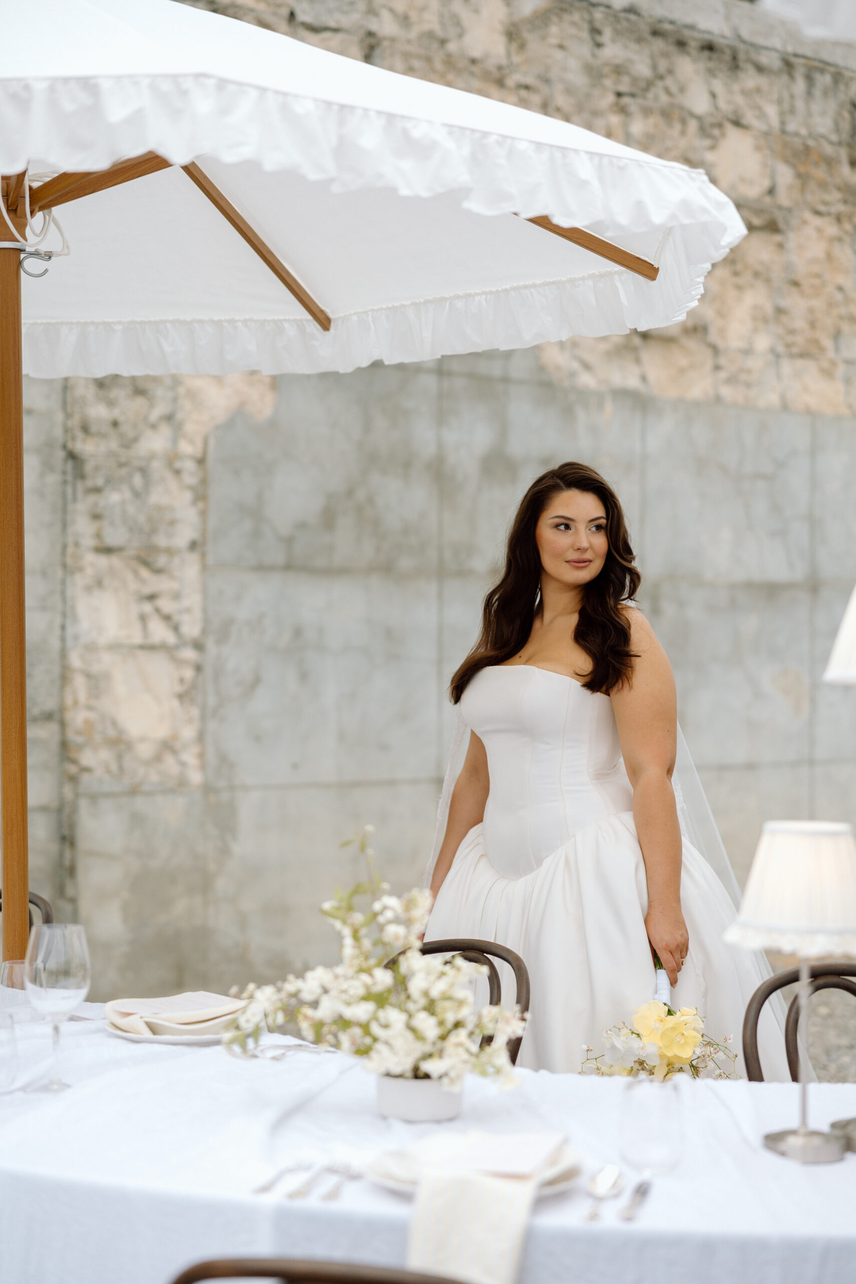 Bride in a strapless satin corset gown with a drop-waist skirt standing behind a styled wedding table, beneath a white ruffled umbrella at The Flaxmill in Oxford, New Zealand